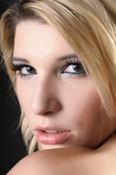 A pretty young woman with blond hair sits in front of a dark background. She is covered only with a white cloth and shows her beautiful,naked back to the camera.