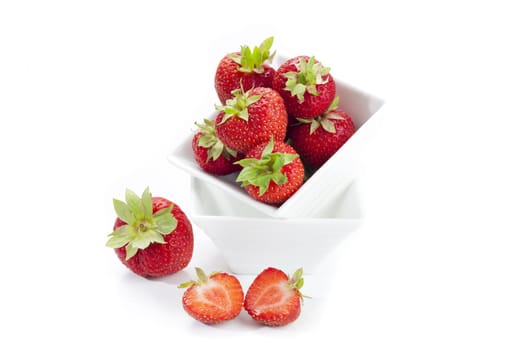 Freshly harvested red strawberry in a white porcelain bowl and two half strawberries in front of it insulated against a white background