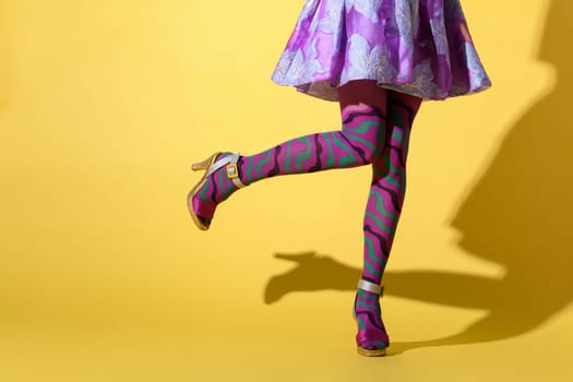 Sexy young woman in flared skirt, purple collant and high heels kicking her foot to the side in a close up on her legs over a yellow studio background with copyspace