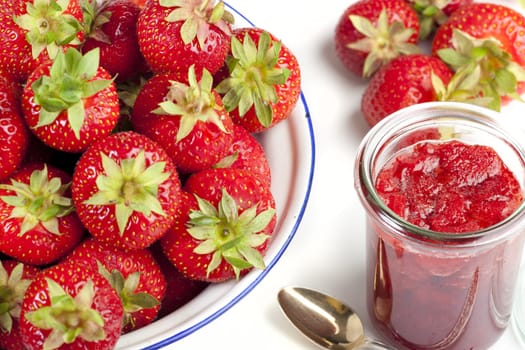 Fresh strawberries in an enamel bowl and a jar of jam with spoon against a white background