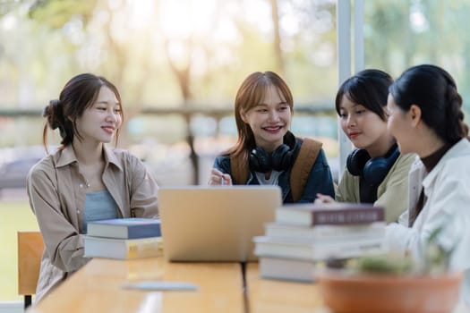 Group of young Asian college students sitting on a bench in a campus relaxation area, talking, sharing ideas, doing homework or tutoring for the exam together.