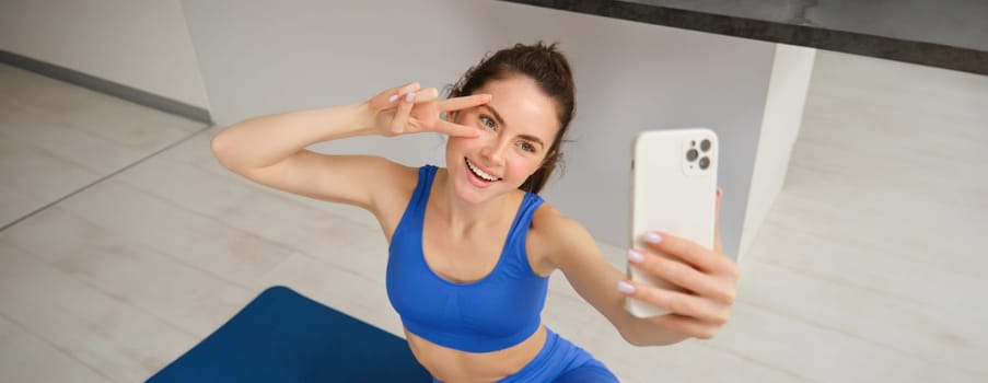 Portrait of young styling fitness girl doing workout from home, taking selfie and video for social media, gym instructor records her training session indoors.