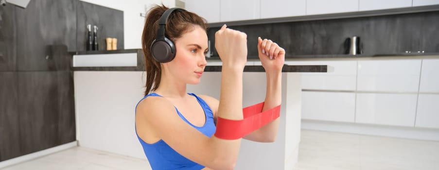 Portrait of determined beautiful woman, listening music, workout with elastic resistance band, doing exercises on arms, wearing sportsbra.