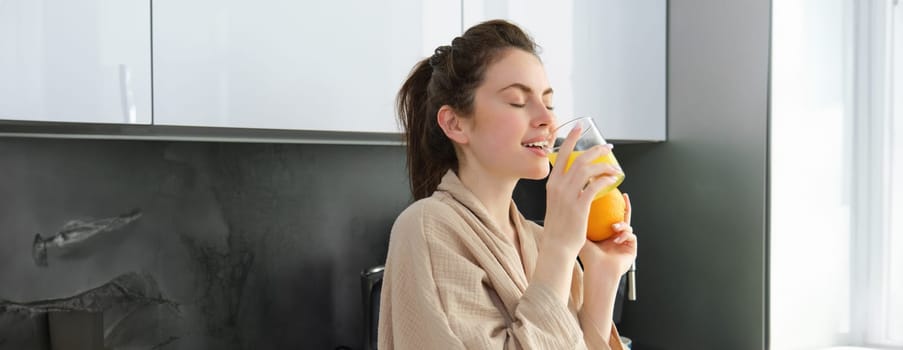 Portrait of happy woman in kitchen, wearing bathrobe, drinking orange juice, freshly squeezed drink, smiling and laughing, food and drink concept.