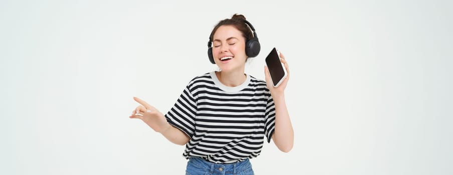 Girl with smartphone dancing, listening to music in wireless headphones, using mobile phone streaming app to enjoy favourite songs, isolated over white background.