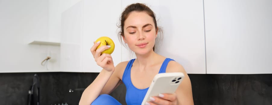 Portrait of sportswoman, girl eating and apple and looking at her social media, smartphone screen, having a snack in kitchen, wearing fitness activewear.