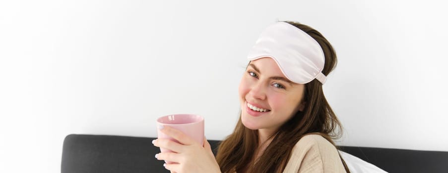 Happy young woman starting her morning with cup of coffee in bed, smiling and enjoying her lazy day, has sleeping mask on forehead.