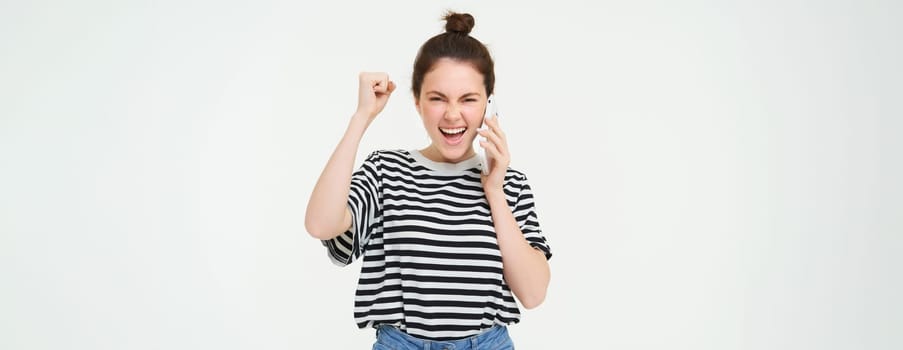 Portrait of enthusiastic young woman, talks on mobile phone, answers phone call and chanting, celebrating, laughing and smiling with excited face expression, white background.