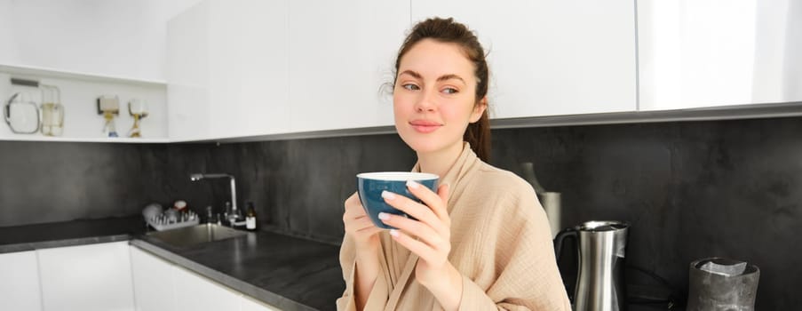 Romantic young woman drinking coffee in kitchen. Girl with mug in hands standing at home wearing a bathrobe, smiling with thoughtful face.
