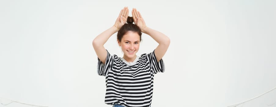 Image of beautiful, happy young woman showing bunny ears on top of her head, looking excited, posing over white background. Lifestyle and people concept