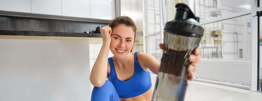 Young positive girl does workout from home, gives you water bottle to drink during training exercises, sits on yoga mat floor in living room.