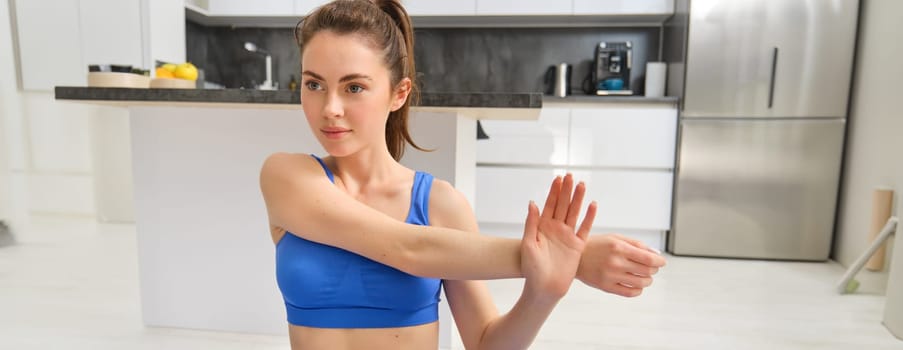 Close up shot of woman doing workout from home, warm-up stretching hands, does fitness exercises in blue sportsbra and smiling.