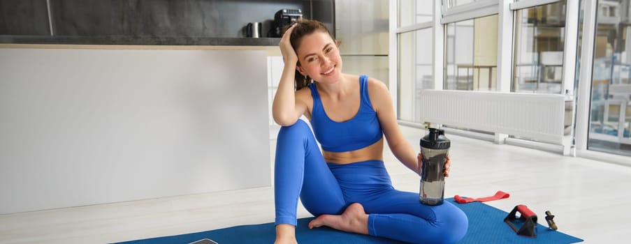 Portrait of beautiful sportswoman in blue sportswear, drinks water, stays hydrated during fitness training session, smiling at camera.