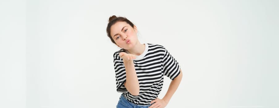 Portrait of beautiful brunette woman sends air kiss at camera, holds palm near lips, isolated against white background.