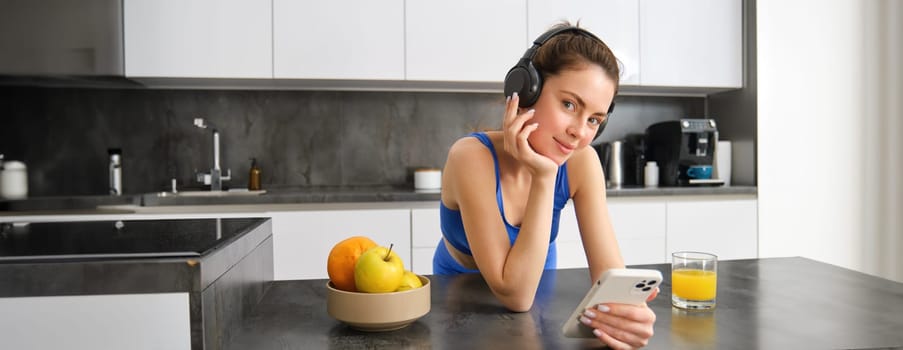 Portrait of young fitness woman with headphones, drinking orange juice in kitchen and using smartphone, listening music, getting ready for workout gym.