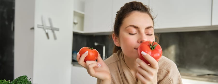 Portrait of beautiful young woman smelling tomatoes, cooking salad from fresh vegetables bought in market, preparing food for family, lead active and healthy lifestyle, standing in kitchen.