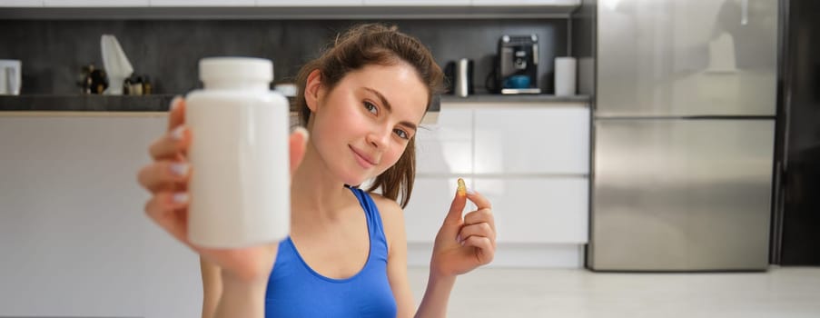 Close up portrait of young woman, fitness instructor showing bottle of vitamins, taking buds supplementary, dietary pill, sitting at home, doing workout exercises.