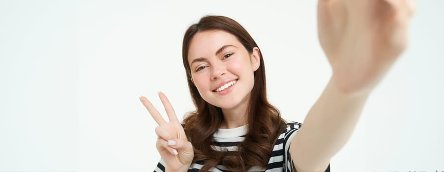 Smartphone view, woman holding mobile phone camera and taking selfie with peace, v-sign gesture, smiling at camera, posing over white background.