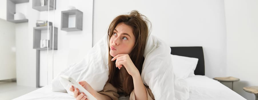 Photo of beautiful joyful woman in pyjama using mobile phone and smiling while lying on bed after sleep or nap.