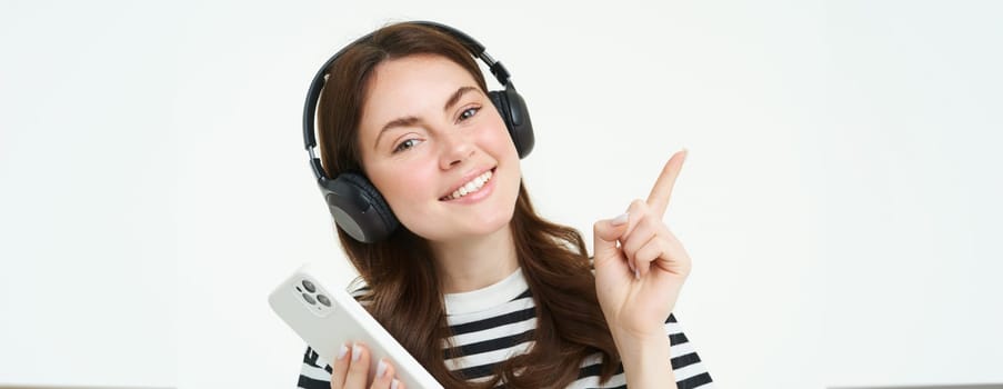 Portrait of smiling student, girl in headphones, holding mobile phone, pointing left, showing advertisement, store offer, white background.