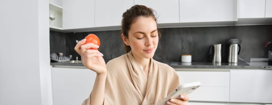Close up portrait of beautiful smiling woman, holding fresh tomato, sitting in kitchen with smartphone, orders vegetables online, using application to buy groceries, using mobile phone.