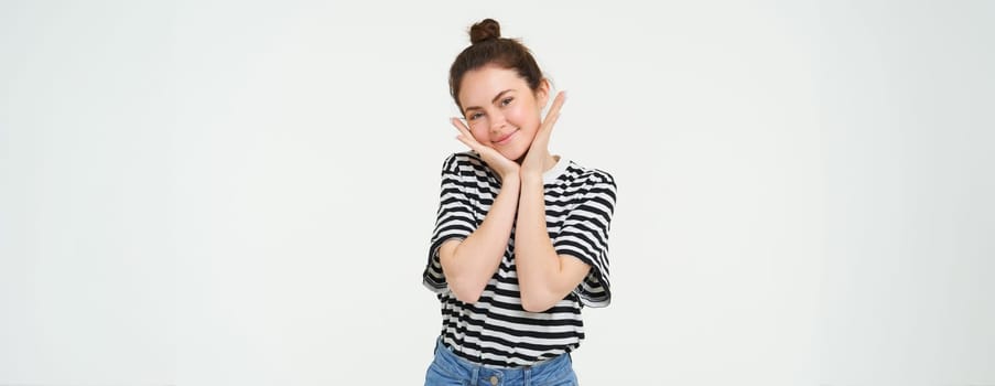 Wellbeing and women concept. Portrait of young woman with cute face, holds hands near head and smiles, isolated against white background.