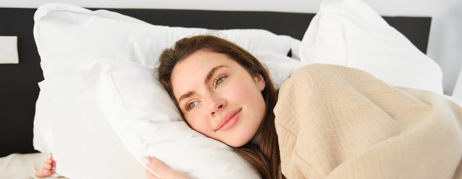 Portrait of happy, beautiful young woman resting in bed, wearing pyjamas, hugging her pillow and smiling while sleeping.
