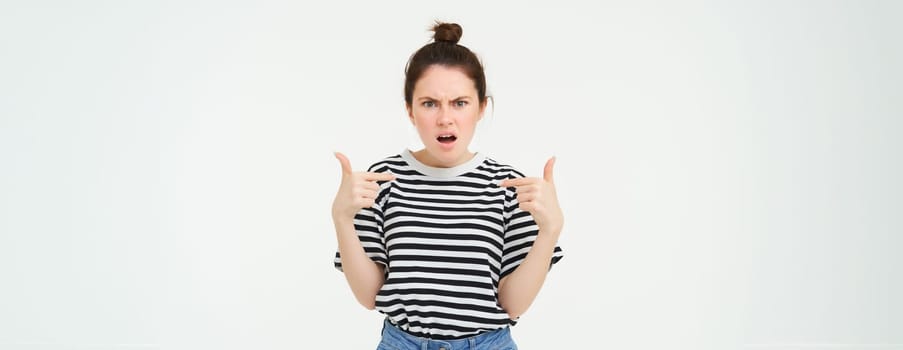 Annoyed woman pointing at herself and frowning, arguing, looking frustrated and disappointed, shouting, standing over white background.