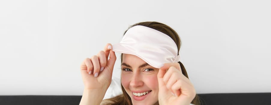 Close up portrait of gorgeous smiling woman with sleeping mask on forehead, waking up in morning, relaxing in bed, laughing and looking happy, resting on vacation in hotel room.