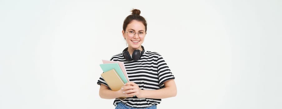 Image of young woman, tutor with books and notebooks, wearing headphones over her neck, isolated on white background. Student lifestyle concept.