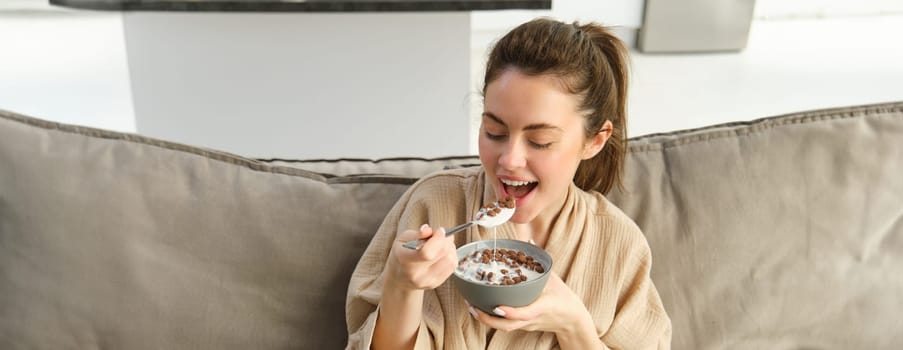 Portrait of gorgeous brunette woman sits on sofa, having her breakfast at home, smiling while eating cereals, holding bowl and spoon and laughing.