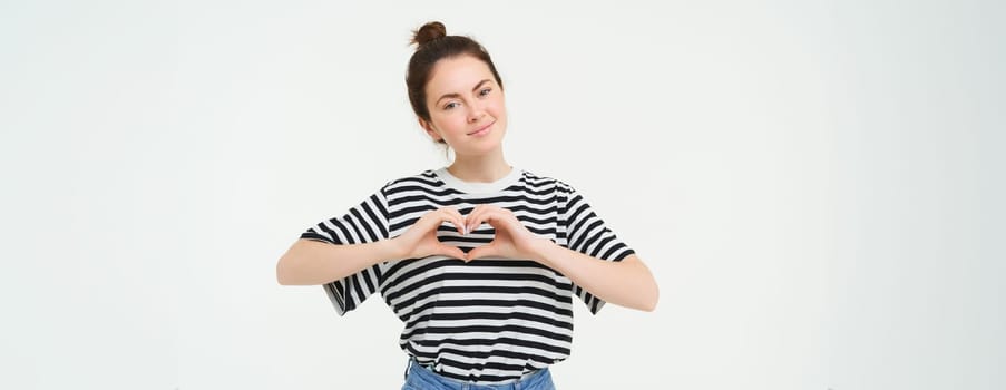 Portrait of beautiful girl shows heart sign, love gesture, like something, stands over white background. Copy space