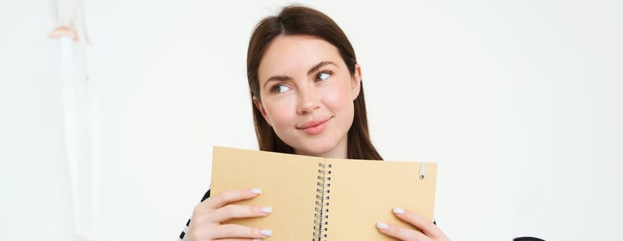 Portrait of beautiful young woman, holding daily planner, writing in her diary, making notes in notebook and smiling, dreaming about something, standing thoughtful against white background.
