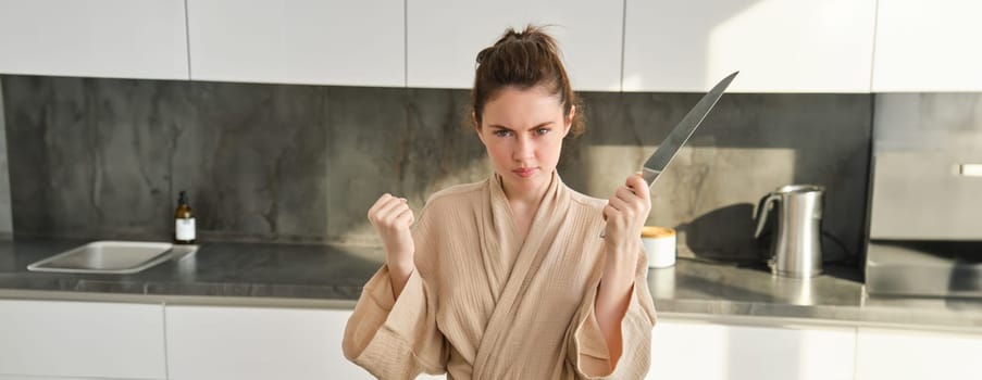 Close up of serious and confident brunette woman holds knife and clenches fist, standing in the kitchen, looking self-assured, posing in bathrobe. Lifestyle and food preparation concept