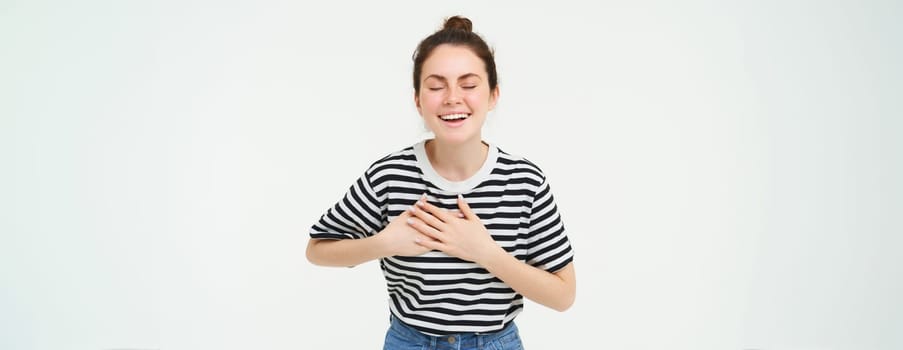 Portrait of happy, smiling young woman stands with eyes closed, holds hands on heart, laughs, remember something funny, isolated over white background.