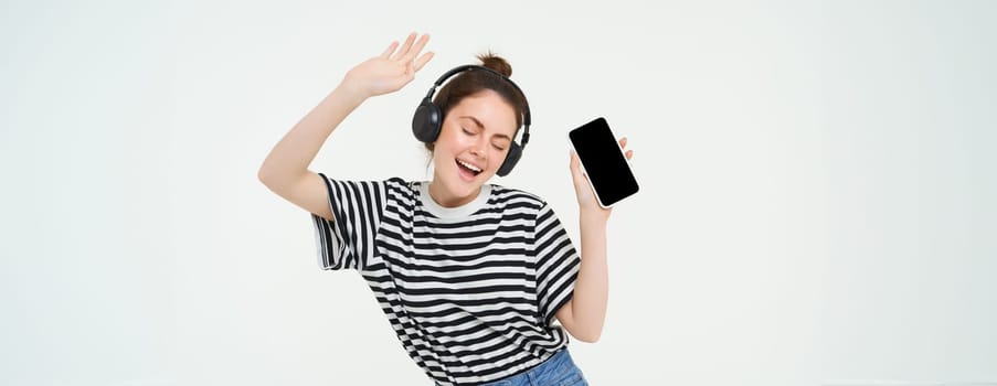 Portrait of stylish young woman dancing in wireless earphones, using headphones and mobile phone music app to listen to favourite songs, posing over white background.