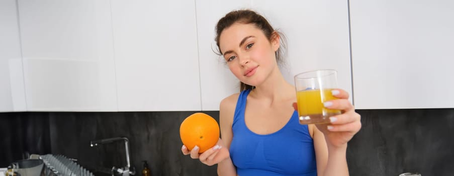 Close up portrait of sportswoman, fitness girl holding glass of fresh juice and an orange in hands, smiling at camera, standing in kitchen. Workout and healthy lifestyle concept