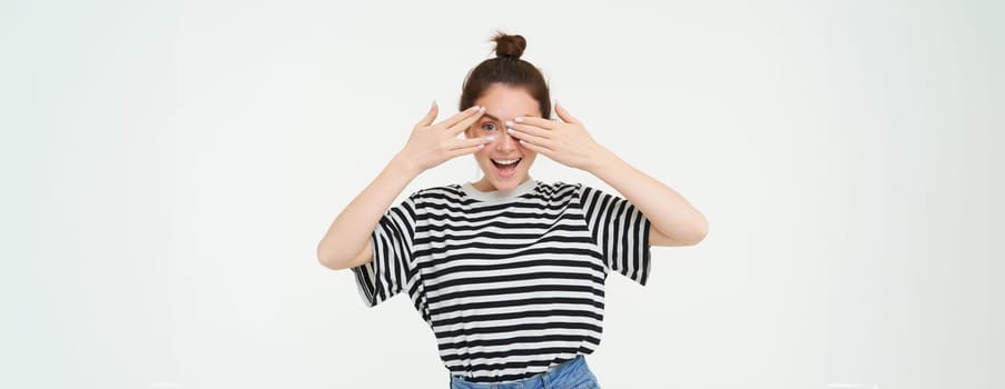 Happy young woman, bday girl peeking through fingers, holds hands on eyes, waiting for surprise, stands over white background.
