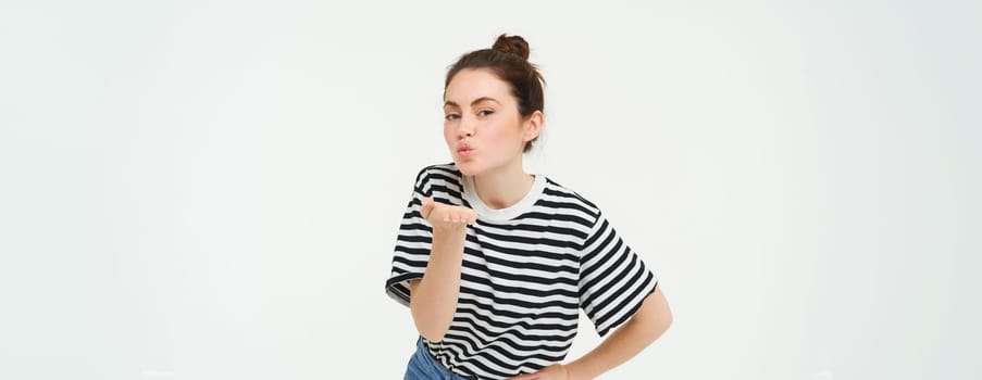 Portrait of cute young woman blows air kiss, sends mwah at camera, posing cute against white background. Lifestyle and people concept.