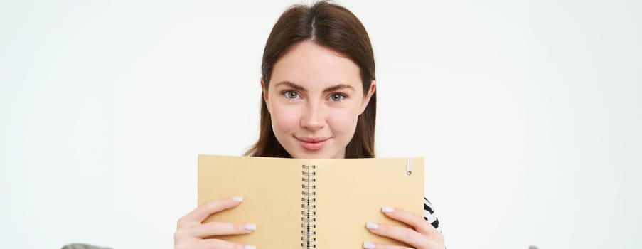 Image of young woman with notebook, holding her personal diary, work planner, smiling, reading something, standing over white background.