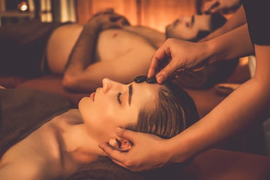 Caucasian couple enjoying relaxing anti-stress head massage with hot stone and pampering facial beauty skin recreation leisure in warm candle light ambient salon spa in resort or hotel. Quiescent