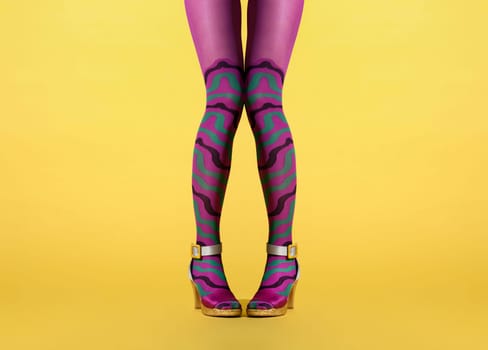 Fun image of the legs of a woman with knock knees and pigeon toes in purple collant or pantyhose decorated with a wavy stripe pattern wearing stylish modern high heel shoes with straps and buckles over yellow