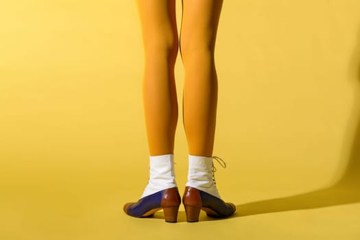 Rear view of a slim woman with skinny legs wearing a mustard yellow collant or pantyhose with retro style tritone leather ankle boots or lace up shoes over a matching yellow studio background