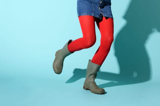 Legs of a woman running in colorful bright red leggings and green rubber boots against a blue studio background with shadow and copyspace