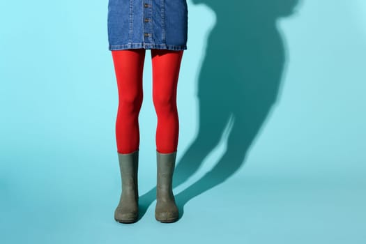 Young woman in green rubber boots or gumboots wearing a short denim skirt and red pantyhose or collant posing over a blue background with copyspace and shadow in a lower body view