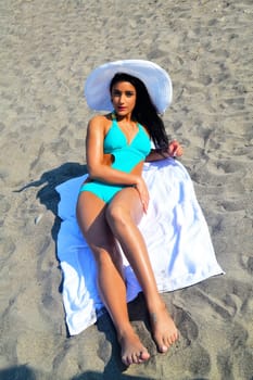 A lovely young woman in a turquoise bikini with a white hat on her black.hair lying on a towel on a sandy beach..