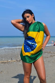A young woman wrapped in a Brazilian flag standing on the beach in bright.sunshine with her long black hair in a closeup shoot...