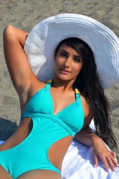 A closeup picture of a beautiful young woman in a turquoise bikini and a.big white sunhat lying on a towel with her long black hair, sunbathing..