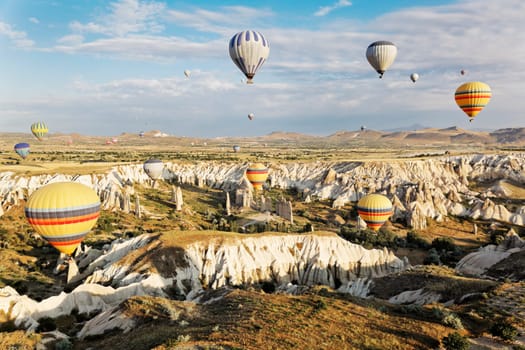 Aerial landscape of earlymorning sun falling of the rough Cappadocia terain whilst overhead the blue skies and clouds are filled with mutli colored hot air balloons