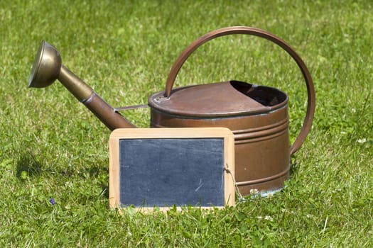 Old copper watering can and an empty slate stand on the green lawn